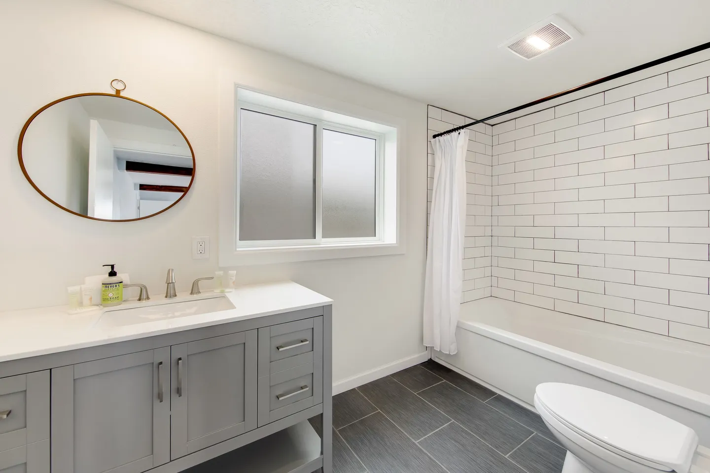 Bathroom with a gray vanity, oval mirror, frosted window, dark tile flooring, and a shower-tub combo with white subway tile.