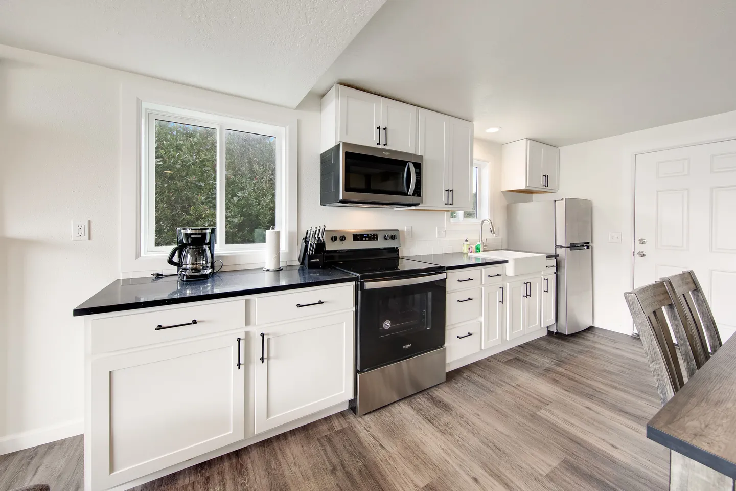Kitchen with white shaker cabinets, black countertops, stainless steel appliances, and wood-look flooring.