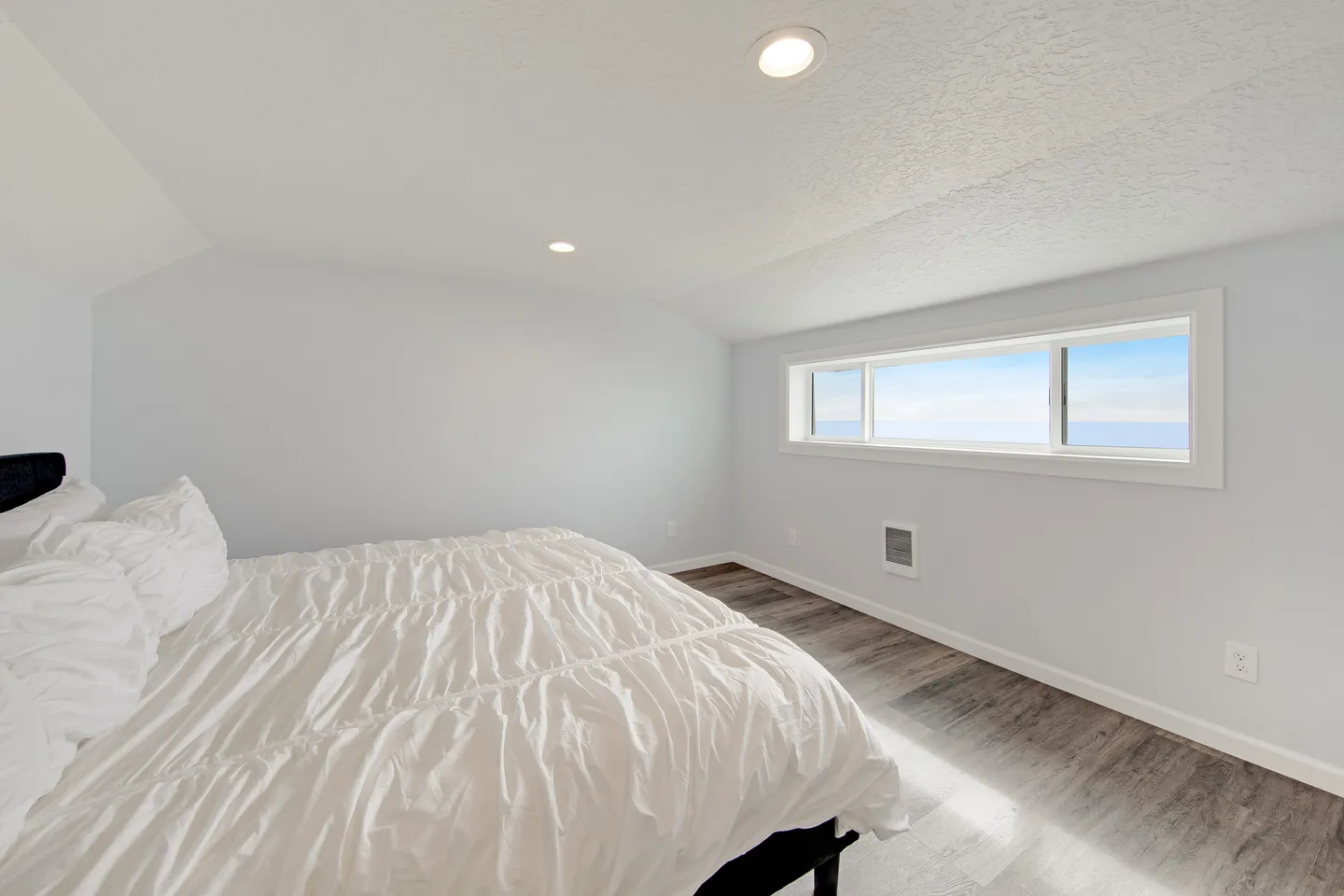 Bedroom with a bed made with white linens, wood-style flooring, white walls, and a horizontal window with a view of the water and sky.