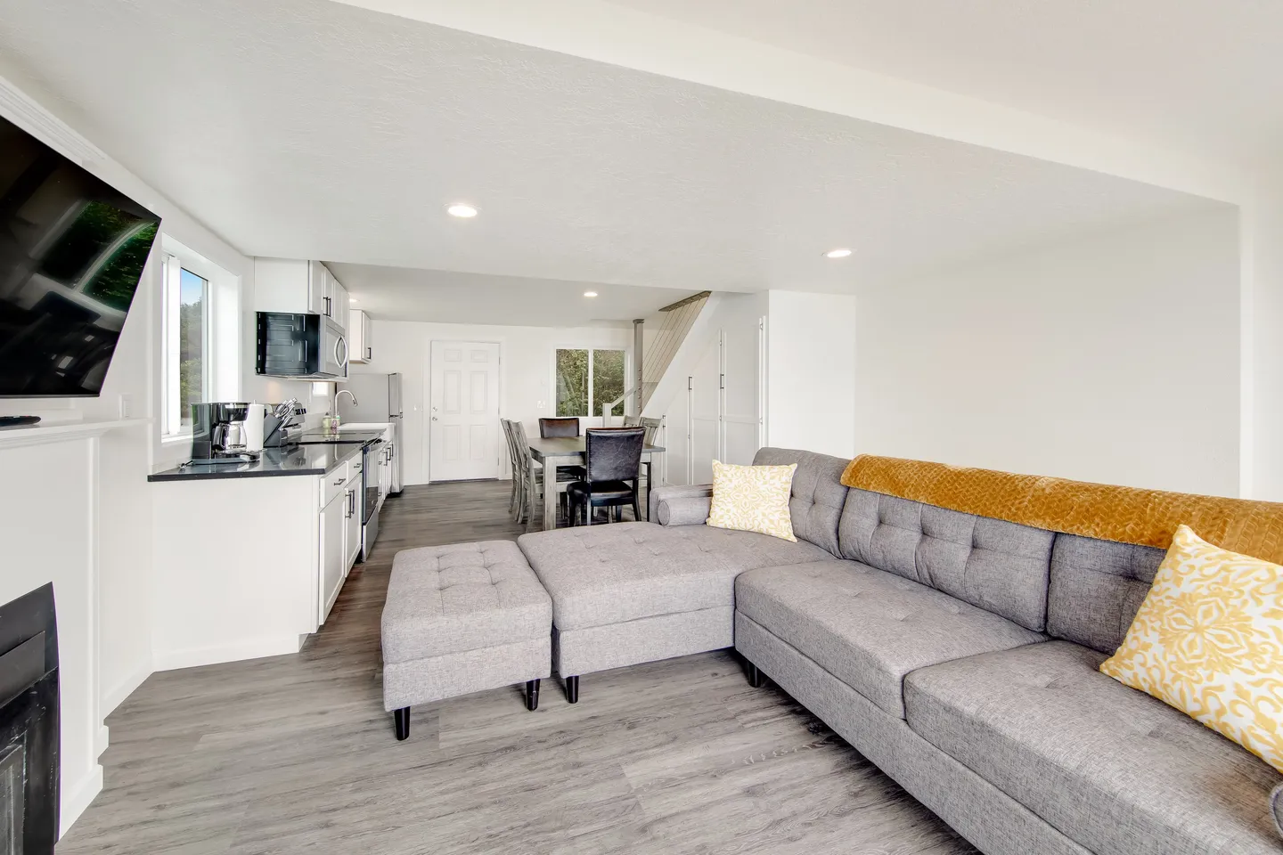 Open-concept living room, kitchen, and dining area with a gray sectional sofa, wood-style flooring, white walls, and a mounted TV.