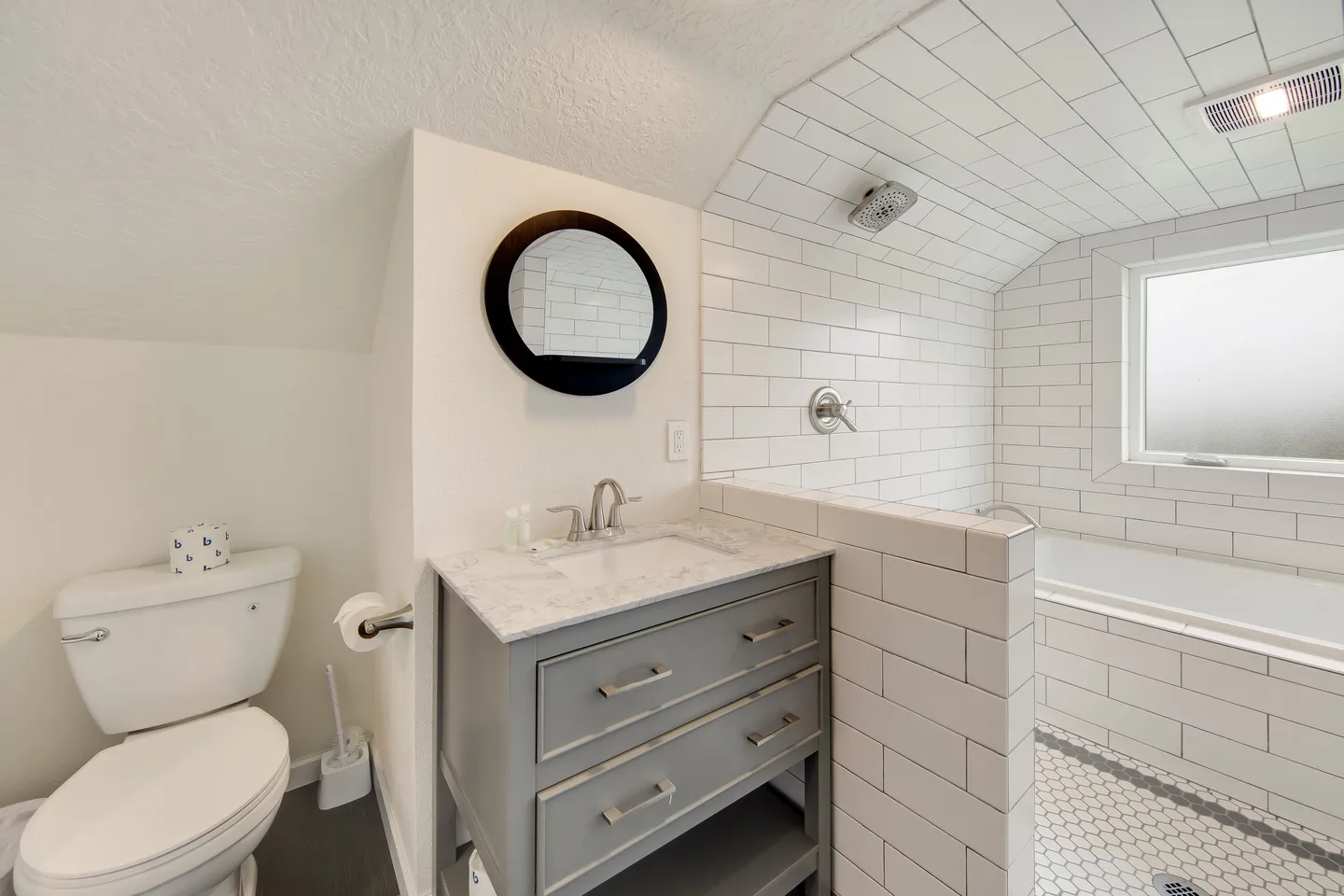 Bathroom with a toilet, gray vanity with round mirror, and a walk-in shower area with subway tile and bathtub.