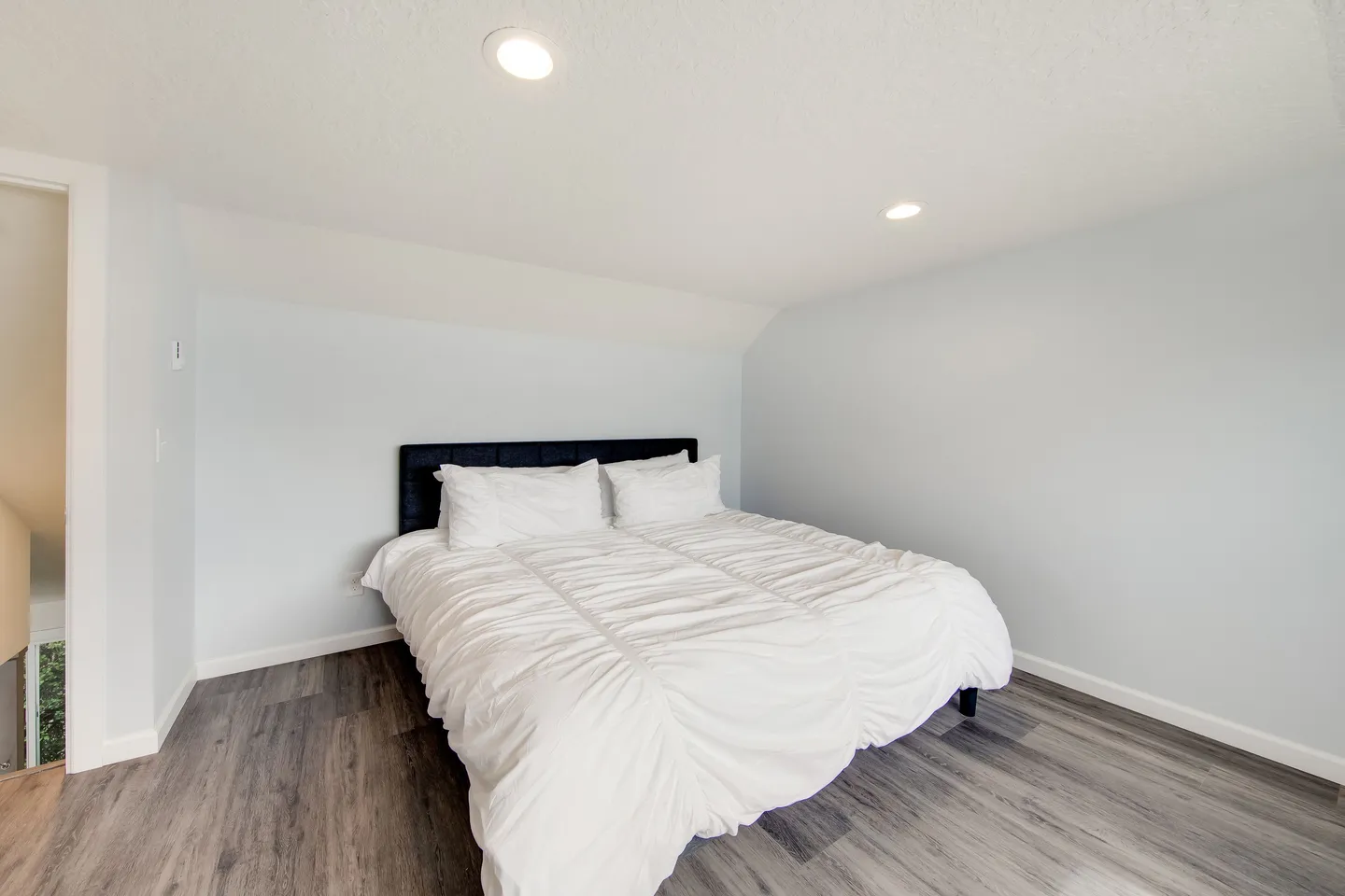 Bedroom with light blue walls, wood-look flooring, recessed ceiling lights, and a bed with a dark headboard and white comforter.