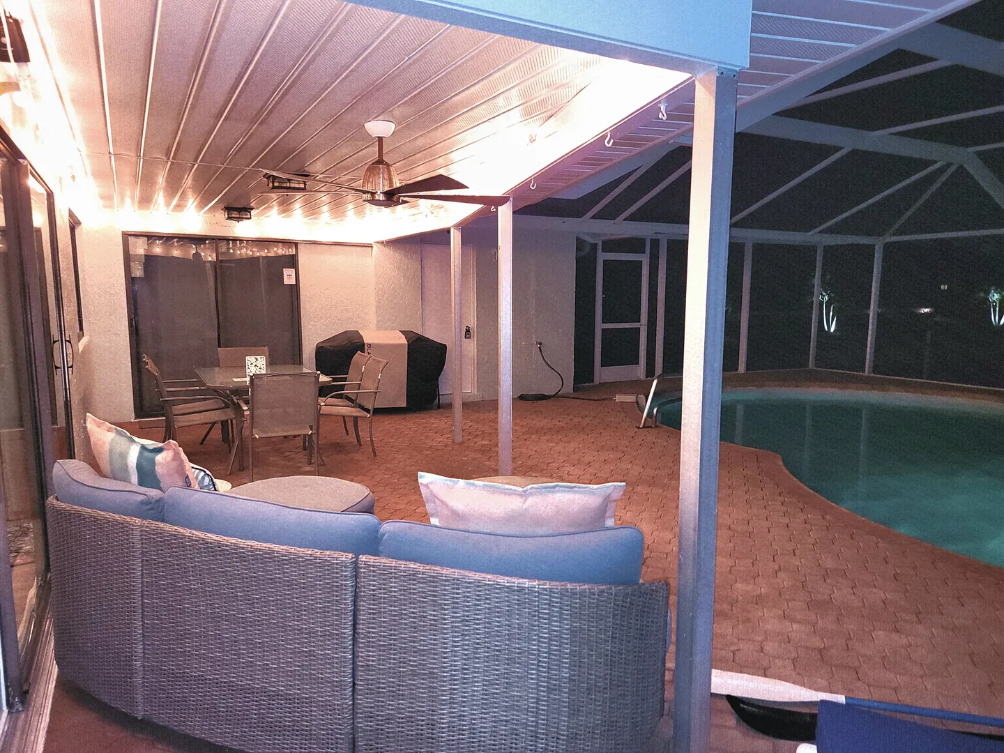 Covered patio at night with woven outdoor sofa, dining table, grill, and swimming pool inside a screen enclosure.