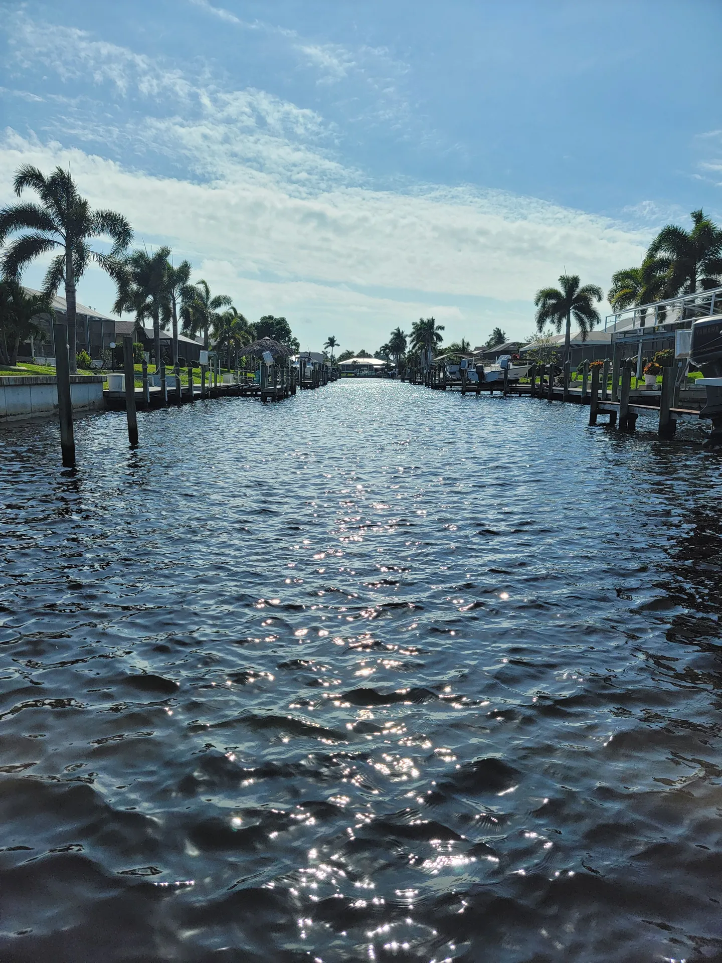 A wide residential canal lined with private boat docks, pilings, and palm trees under a partly cloudy sky.
