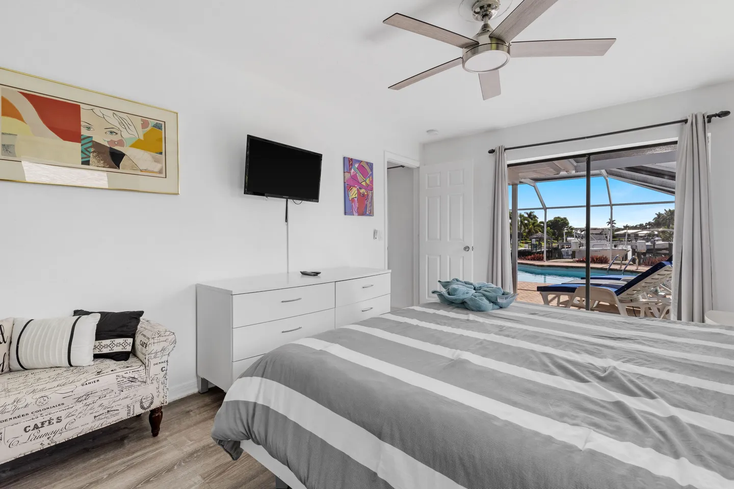 Bedroom with striped bedding, a white dresser with a wall-mounted TV, an accent armchair, and sliding glass doors opening to a pool patio.