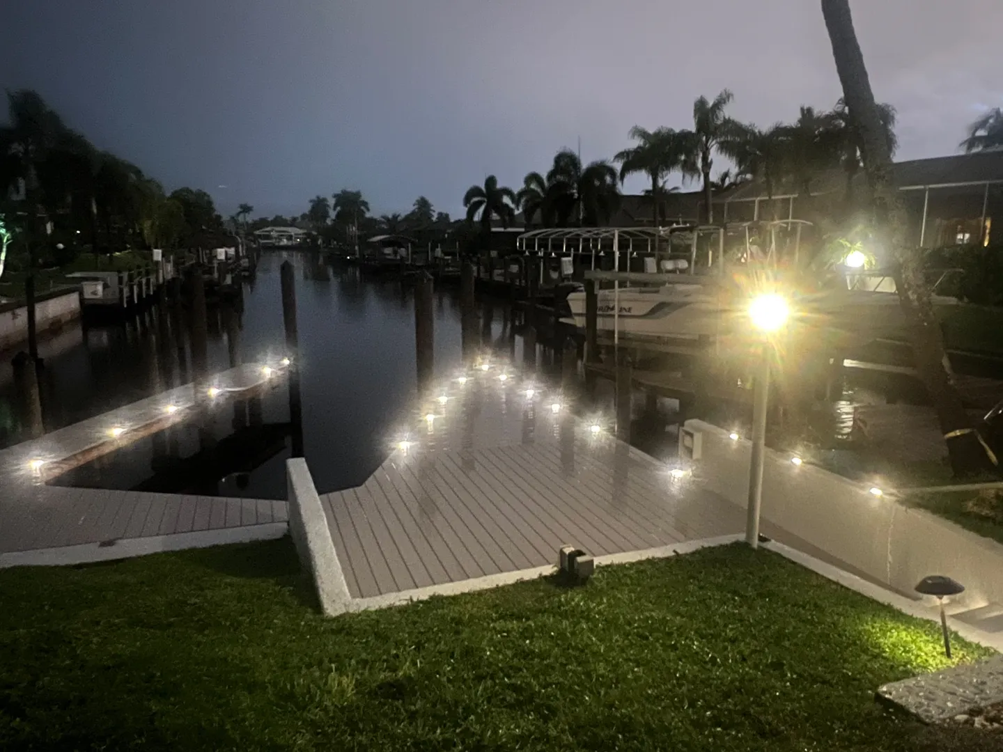 Nighttime view of a boat dock with glowing deck lights along a canal with palm trees in the background.