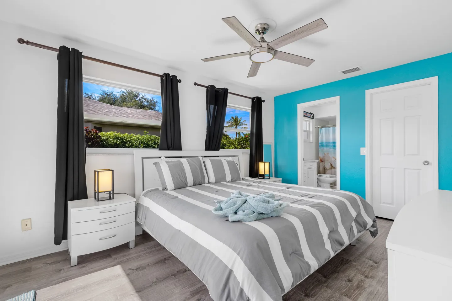 Bedroom with a striped bedspread, white nightstands, ceiling fan, and a turquoise accent wall leading to an ensuite bathroom.