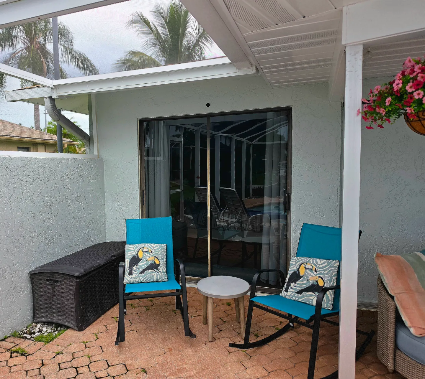 Covered outdoor patio with brick pavers, two bright blue rocking chairs, a small table, and sliding glass doors.