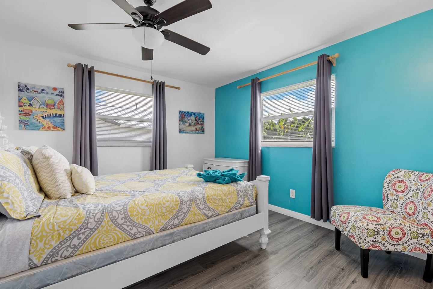 Bedroom with a white wooden bed frame, vibrant turquoise accent wall, patterned armchair, and ceiling fan.