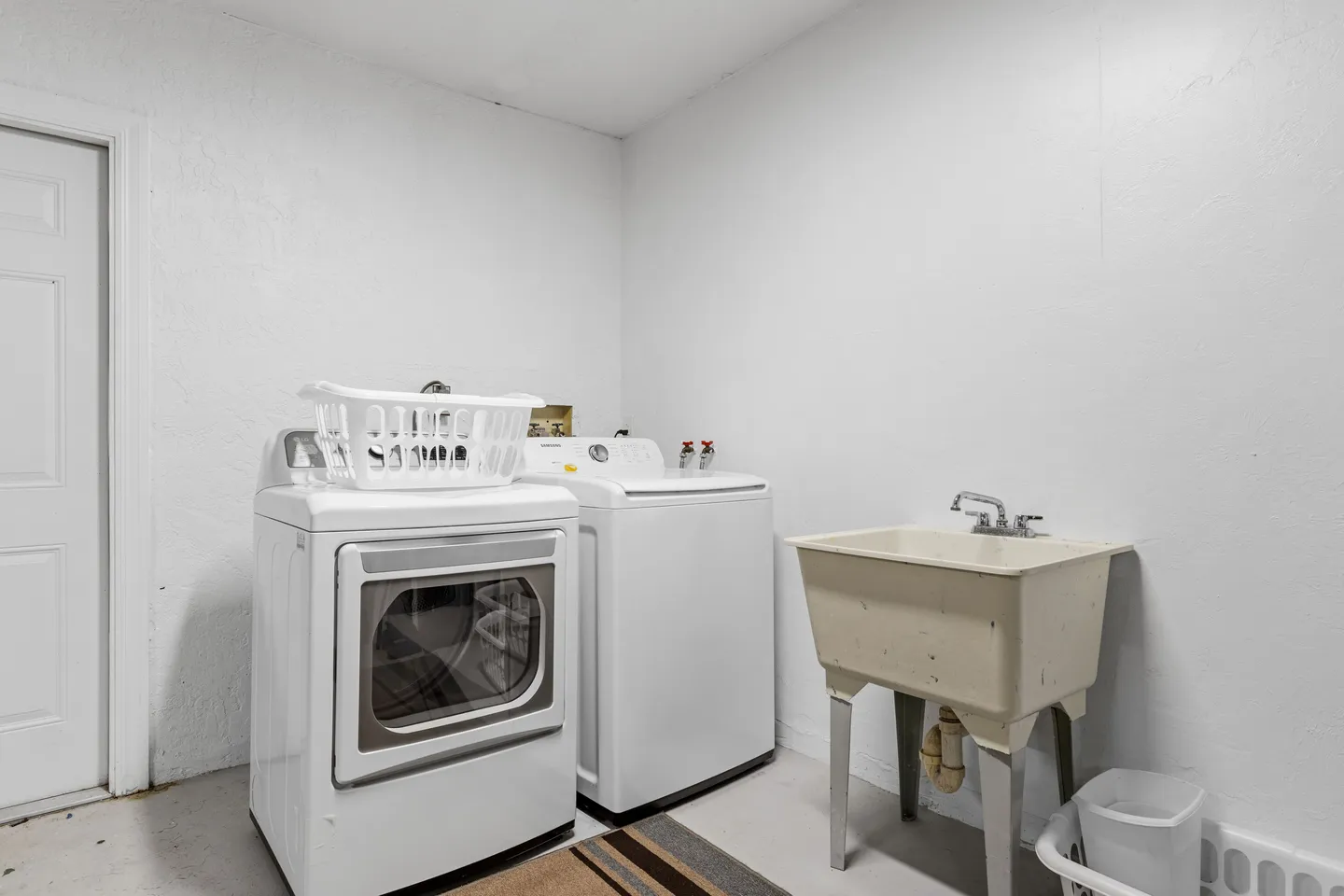 A utility room with a white front-load dryer, top-load washing machine, and freestanding utility sink against white walls.