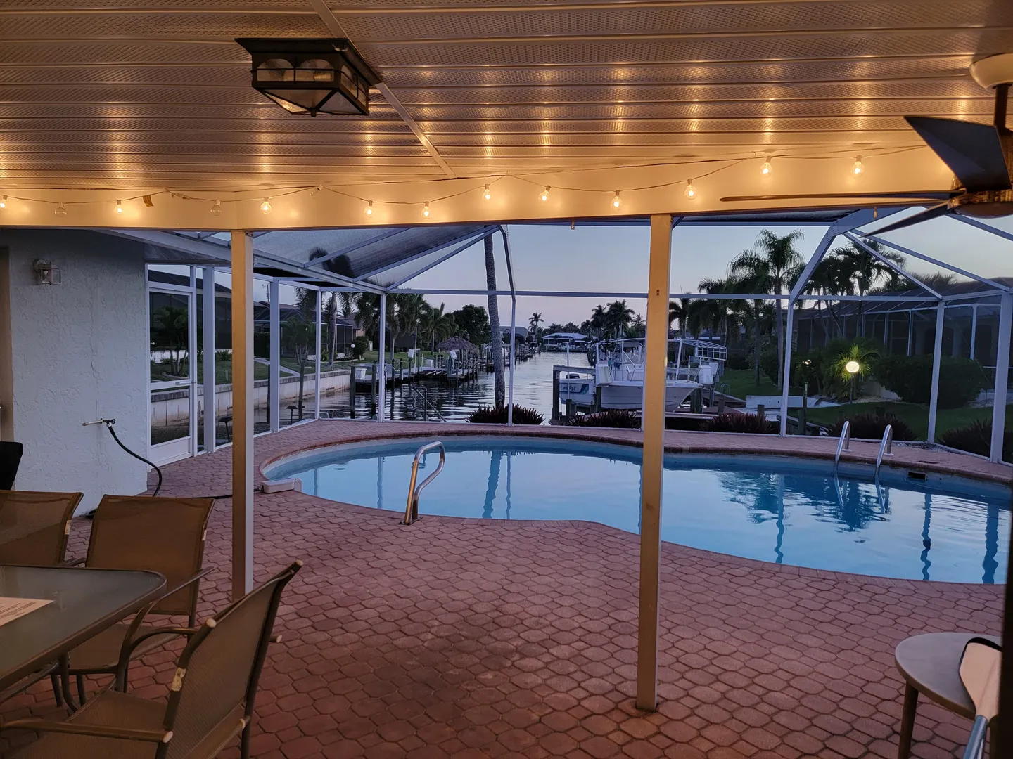 A covered patio with string lights looking out over a swimming pool within a screened enclosure toward a canal with boats at dusk.