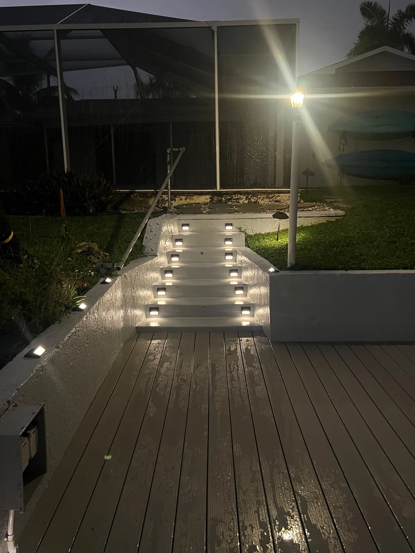 Outdoor illuminated steps leading down from a yard to a wet wooden deck at night.