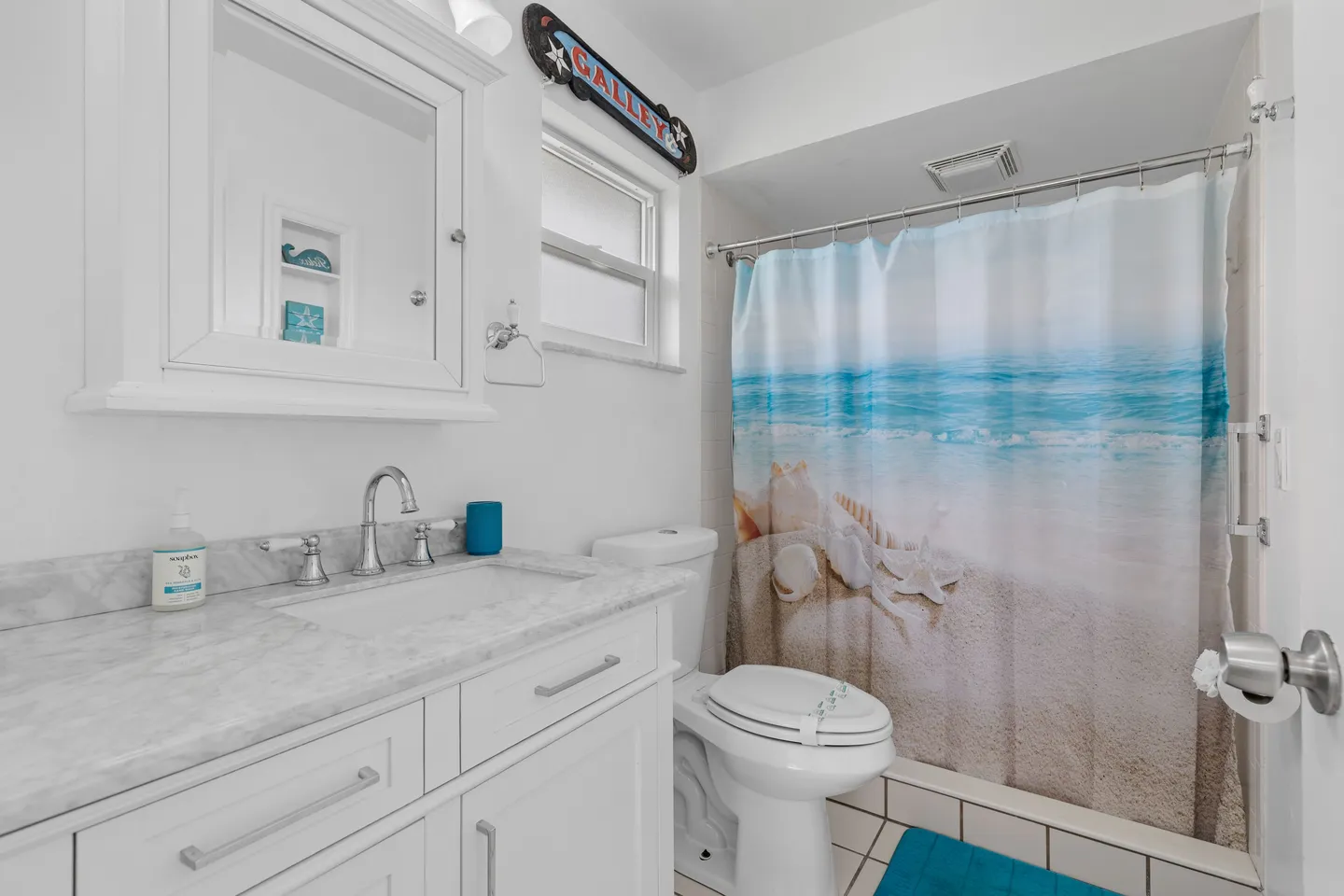 Bathroom featuring a white vanity with marble-patterned countertop, medicine cabinet, toilet, and beach-themed shower curtain.