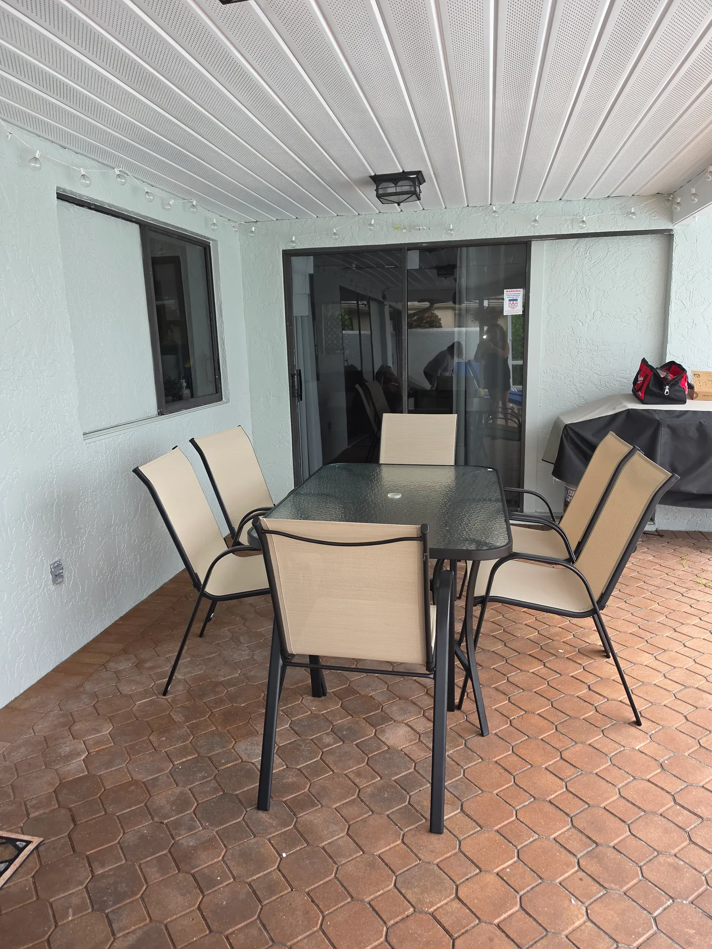 A covered patio with a paver floor, an outdoor dining table, and six chairs near sliding glass doors.