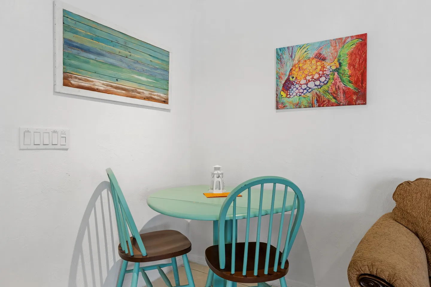 A small circular mint-green table with two matching spindle-back chairs set against a white corner wall with colorful wall art.