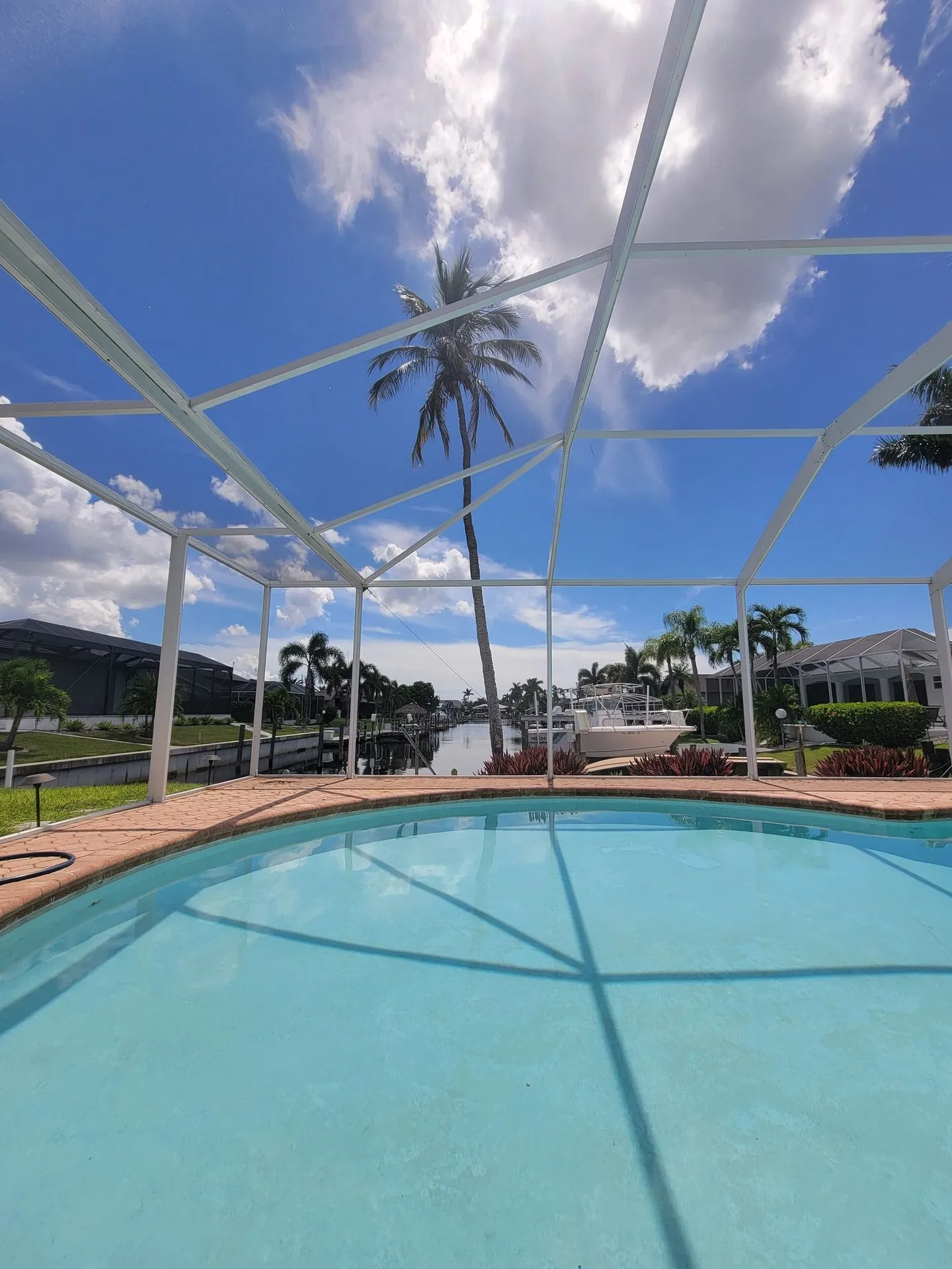 Swimming pool under a white screened enclosure overlooking a canal with palm trees and boats.