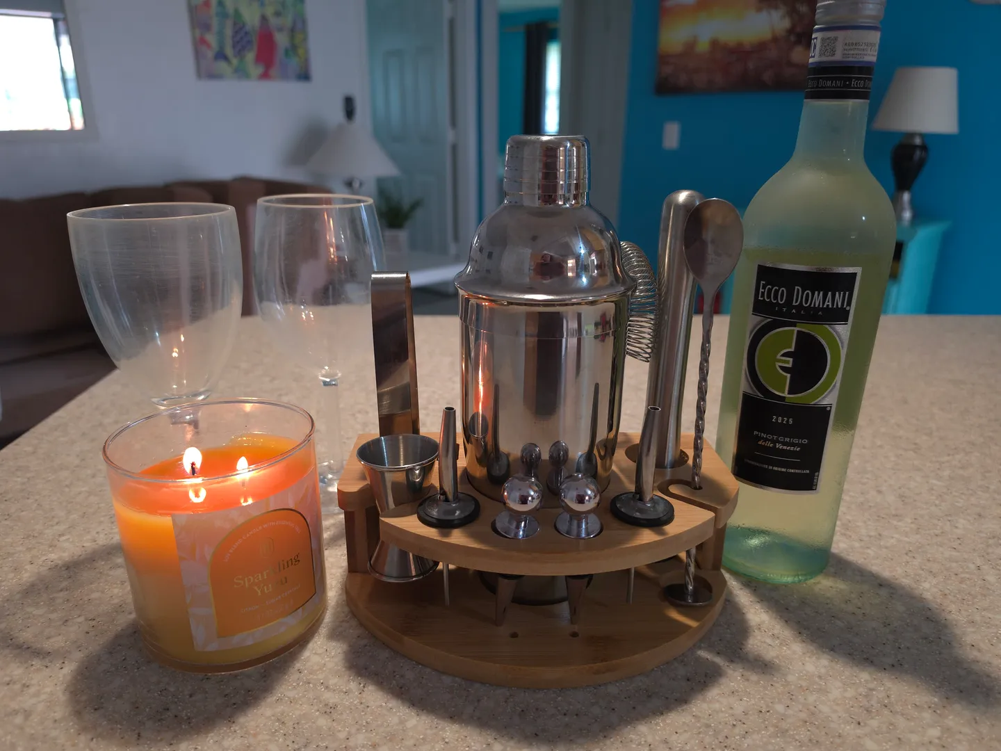 Close-up of a cocktail shaker set on a wooden stand, a bottle of white wine, two glasses, and a lit candle on a counter.