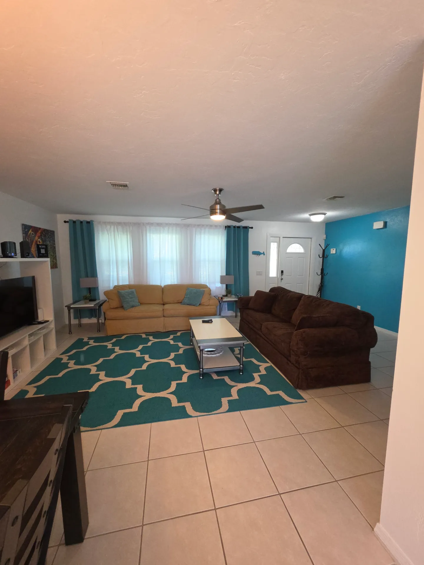 Living room with tiled floors, a yellow sofa, a brown sofa, a turquoise patterned rug, and a turquoise accent wall.