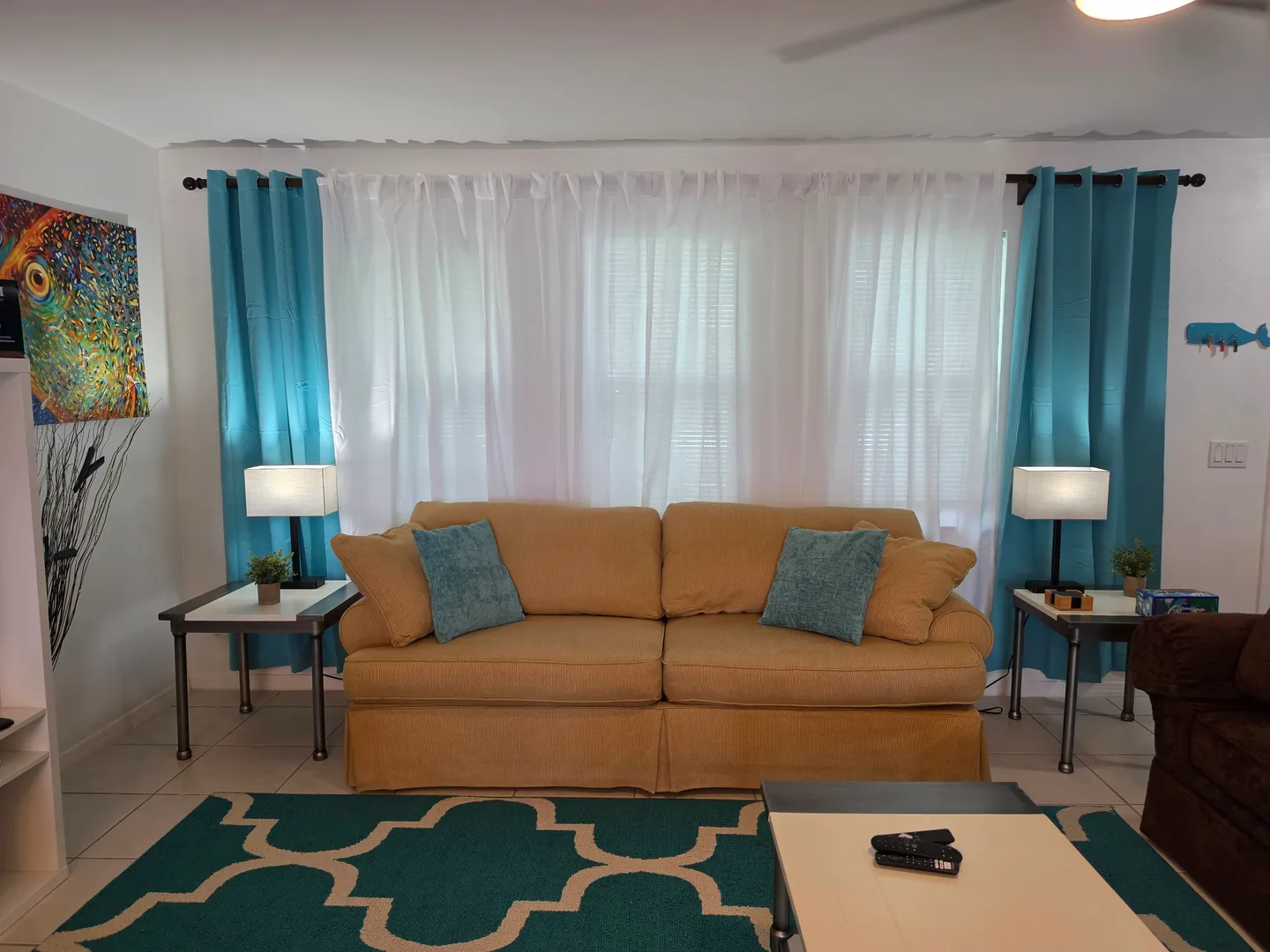 Living area featuring a tan sofa flanked by two end tables with lamps in front of a window with white and teal curtains.