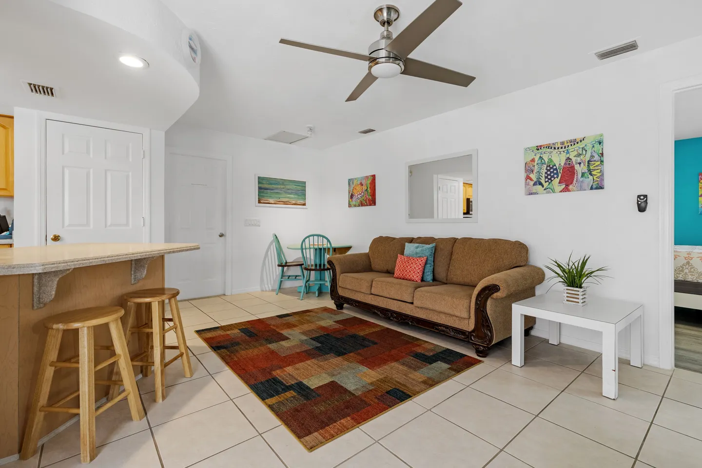 Living area with tiled floors, a brown sofa, a patterned area rug, a ceiling fan, and bar stools next to a counter.