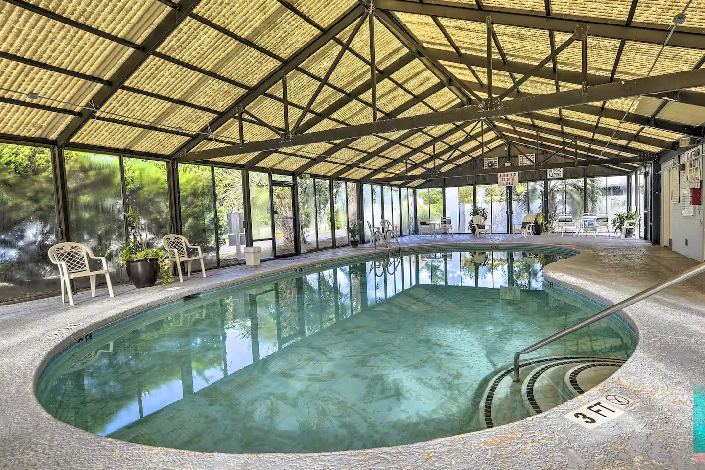 Oval swimming pool inside an enclosed structure with large screened walls and metal truss roof framing.