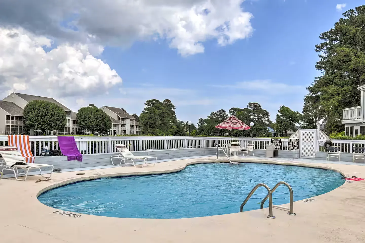 Outdoor curved swimming pool surrounded by a concrete deck with lounge chairs and a white railing fence.