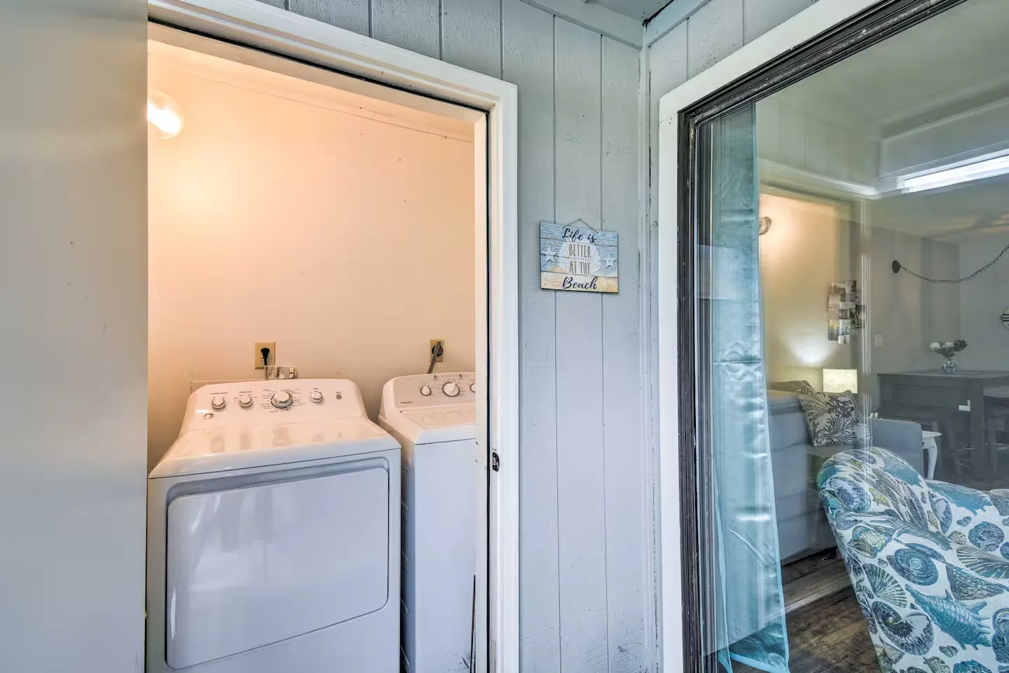 Outdoor laundry closet with washer and dryer next to a sliding glass door looking into a living room.