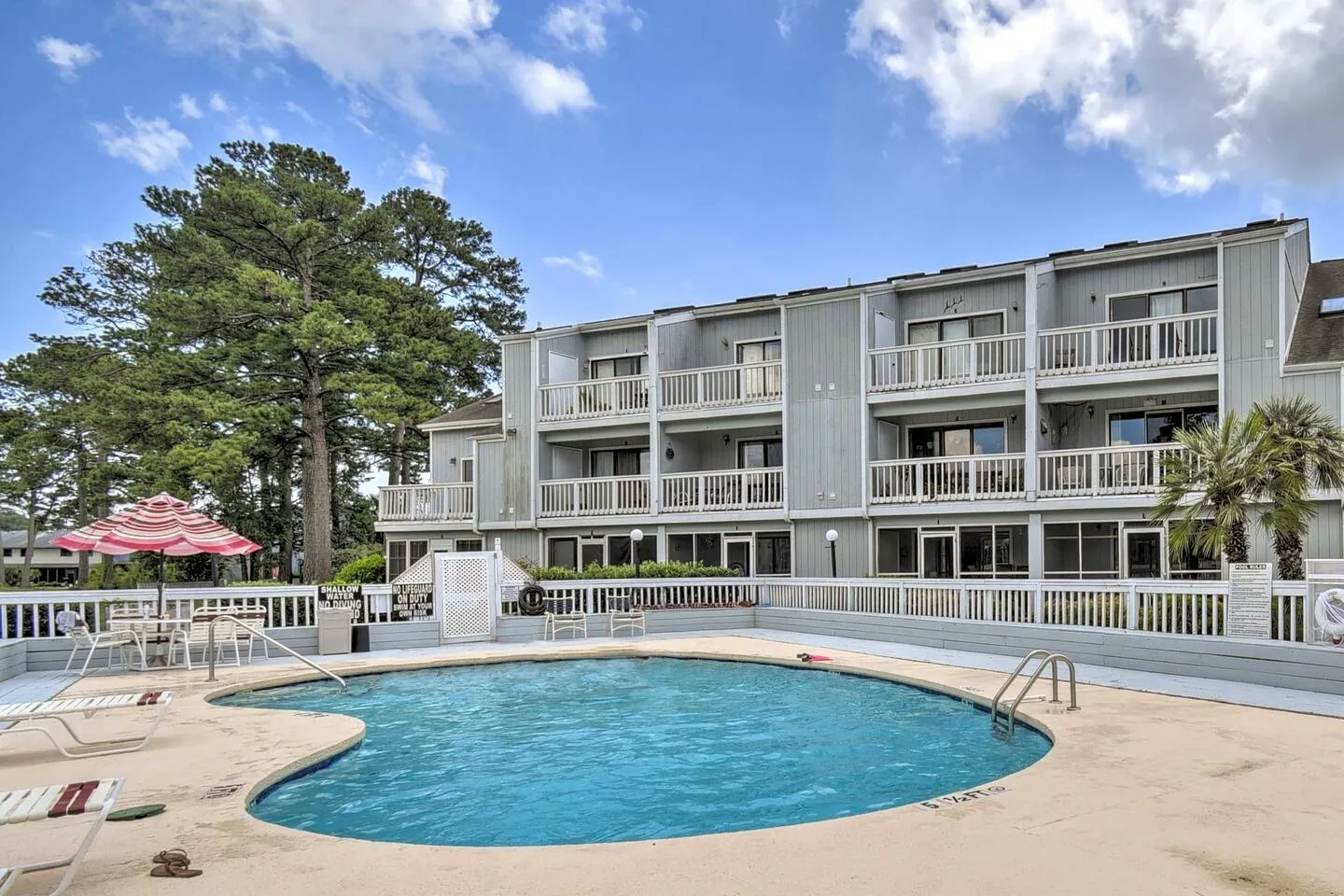Outdoor swimming pool surrounded by a concrete patio, white fence, and a multi-story gray residential building.