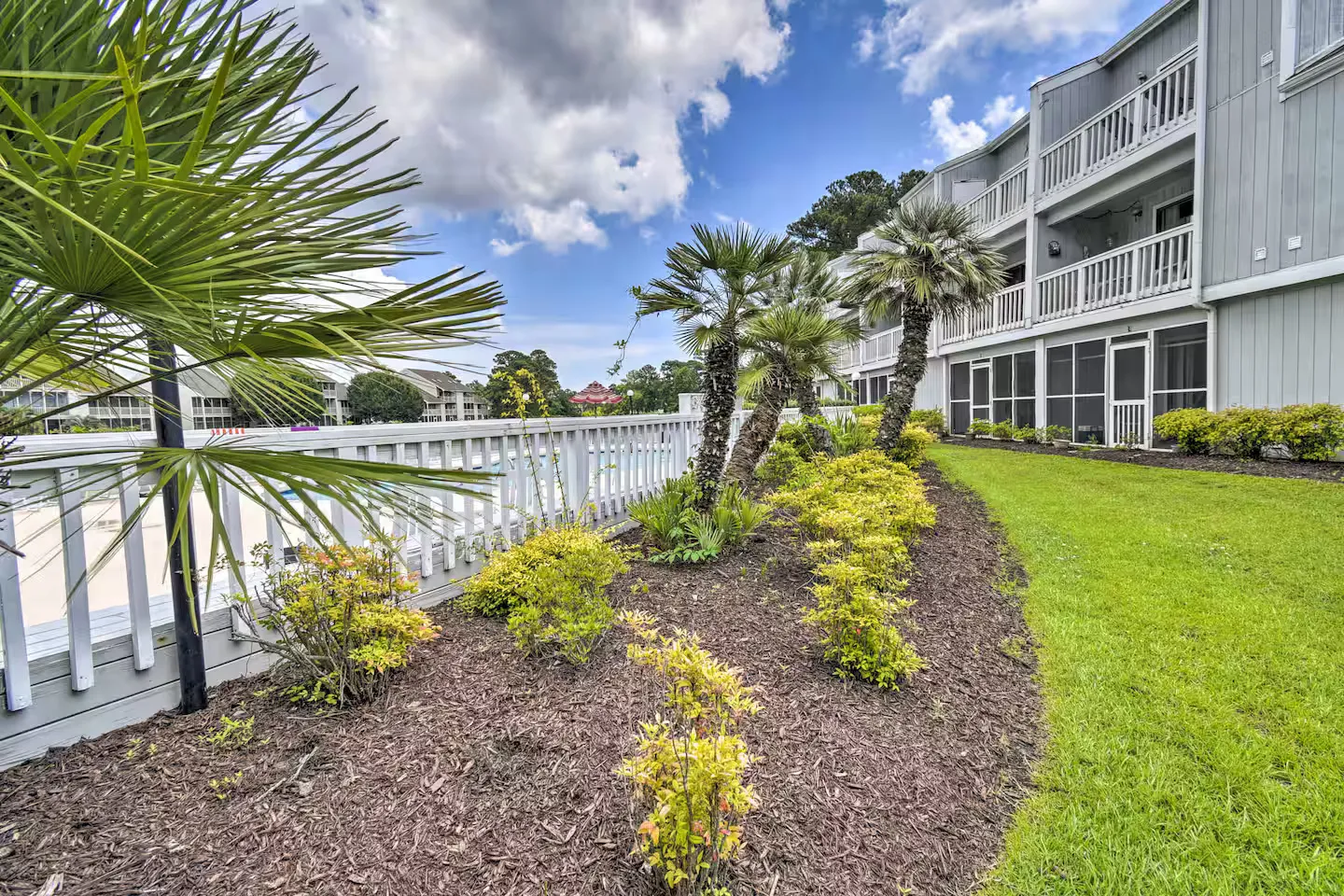 Exterior view of a multi-story residential building with screened porches, a landscaped mulch bed with palm trees, and a white deck railing under a partly cloudy sky.