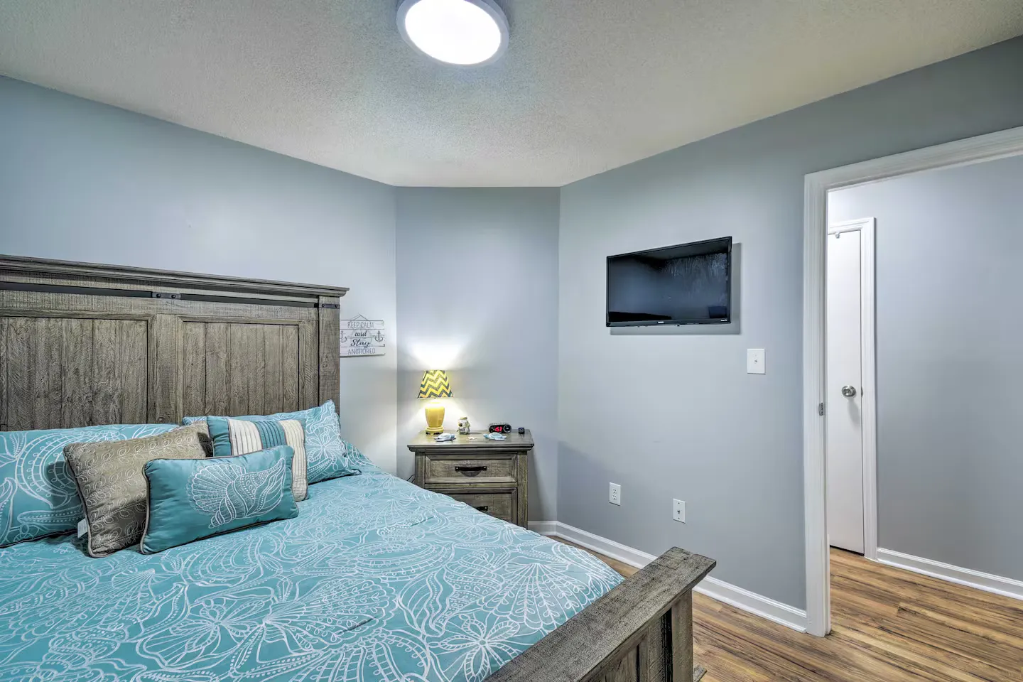 Bedroom with blue walls, wood-finish bed frame, teal patterned bedding, small nightstand, and wall-mounted TV.