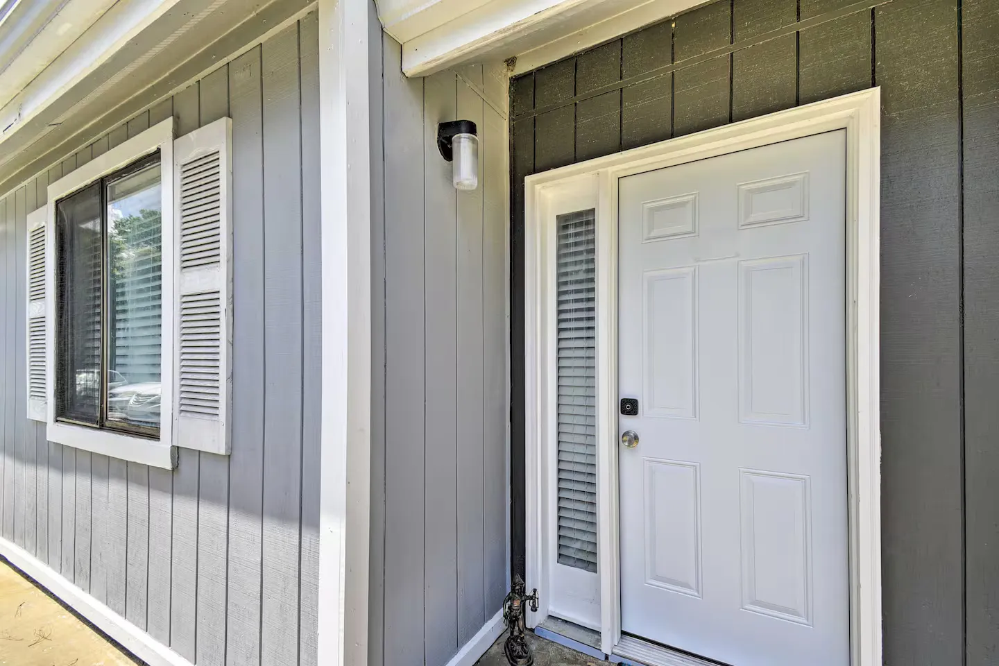 Exterior entryway featuring a white panel front door with a sidelight window alongside grey vertical siding and a shuttered window.