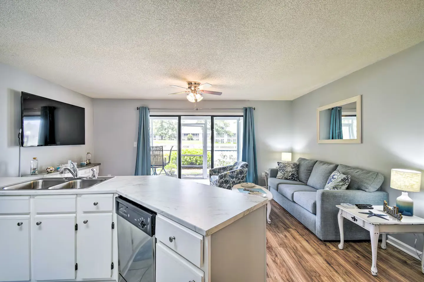 Open-concept living room and kitchen featuring a marble-look peninsula, gray sofa, wall-mounted TV, and sliding doors to an outdoor patio.