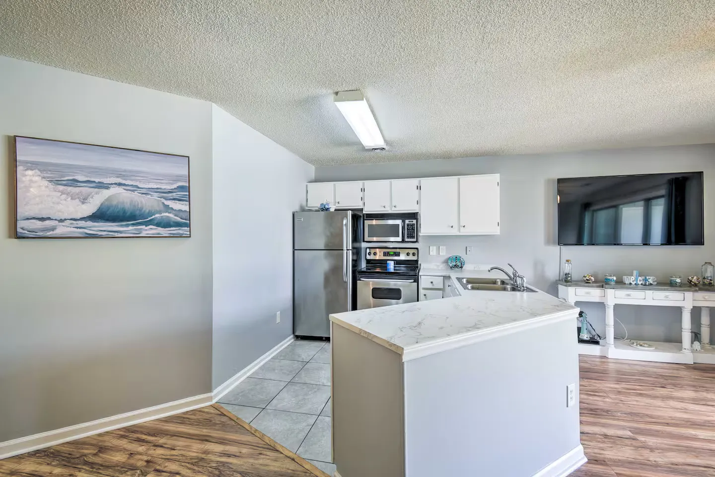 Kitchen with white cabinetry, stainless steel appliances, and a marble-look countertop island, opening to an adjoining room with wood-style flooring and a wall-mounted TV.