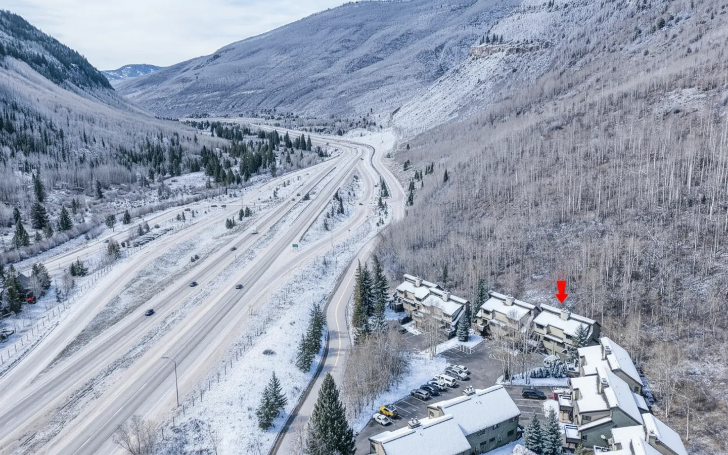 Aerial view of a snow-covered mountain valley with a multi-lane highway, forested slopes, and a residential complex marked with a red arrow.