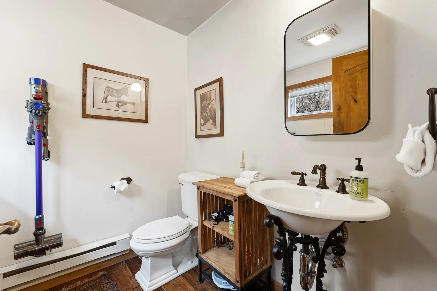 Bathroom featuring a white toilet, a vintage-style pedestal sink with dark ornate legs, a wood side table, and a wall-mounted cordless vacuum.