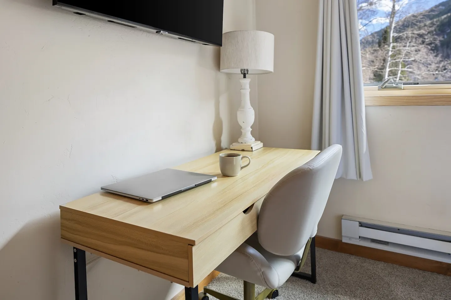 A light wood desk with a laptop, mug, lamp, and chair set against a wall next to a window.