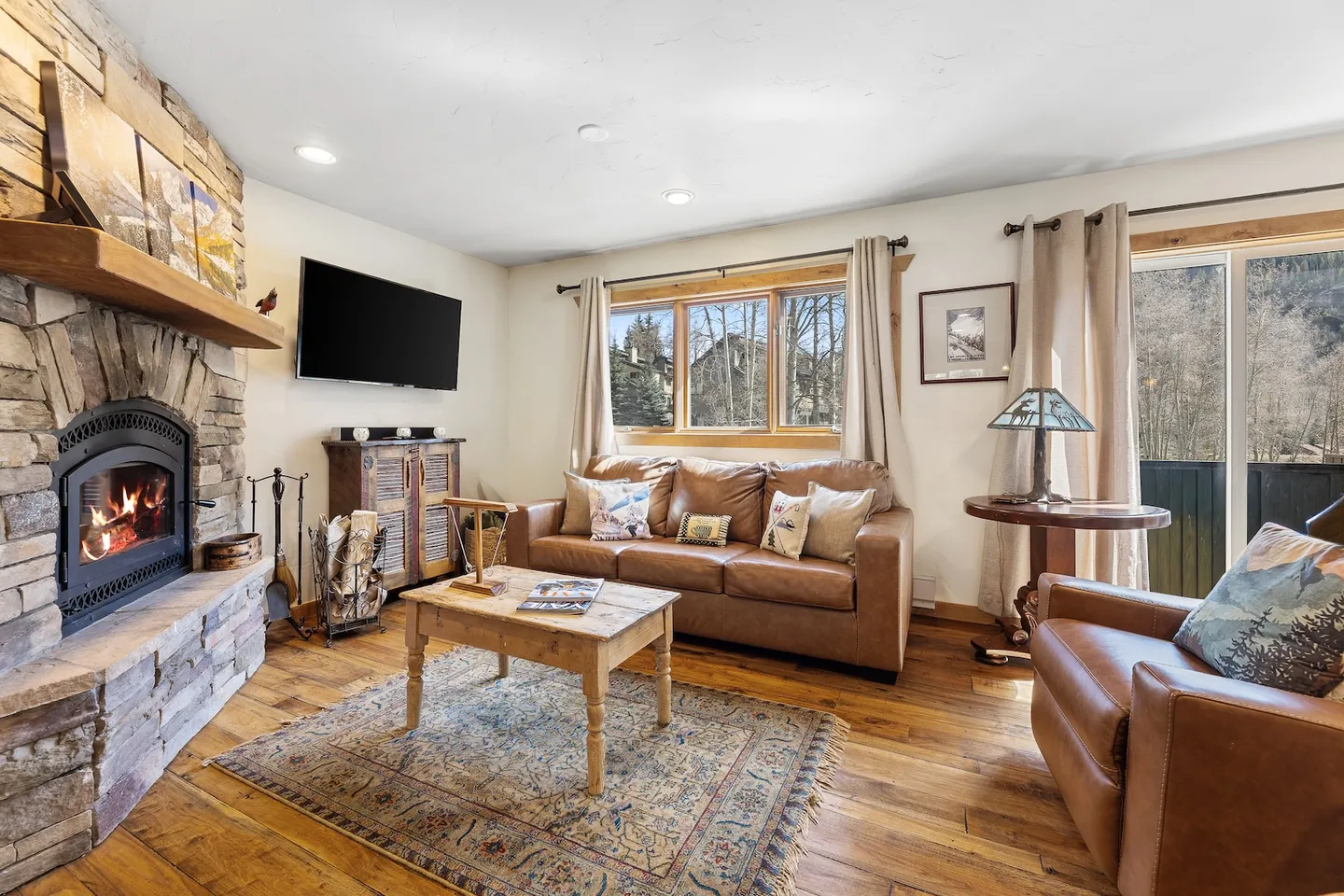 Living room with leather seating, wood-burning stone fireplace, wall-mounted television, and wood floors.
