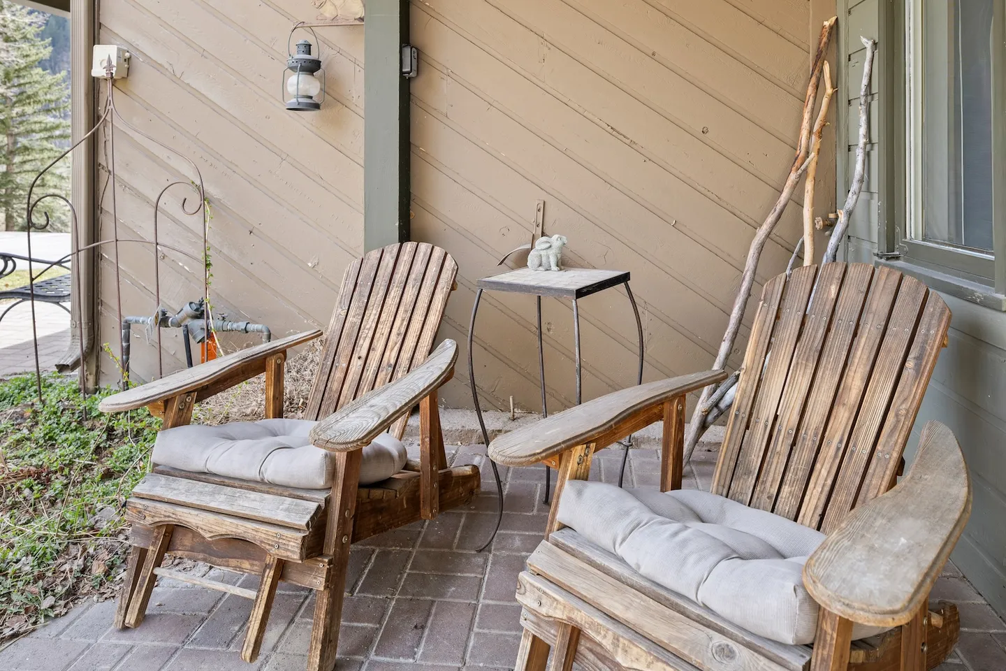 Two rustic wooden outdoor armchairs with seat cushions positioned on a brick patio alongside the wood siding of a house.