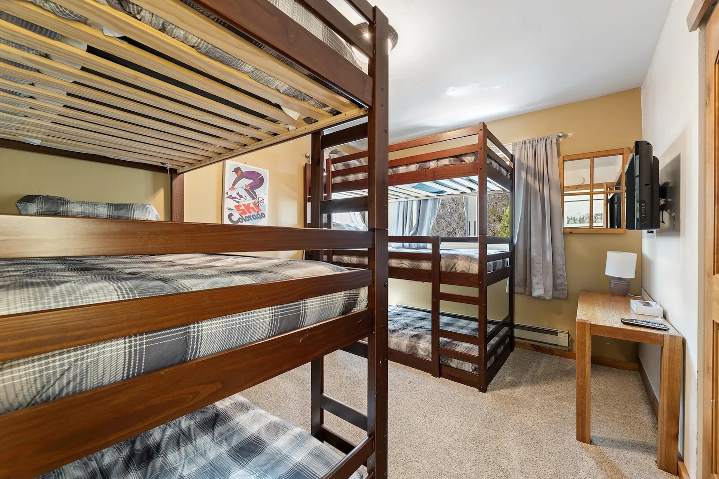 Bunk bedroom featuring multi-tiered dark wood bunk beds with plaid bedding, carpeted floors, and a small desk with a lamp.