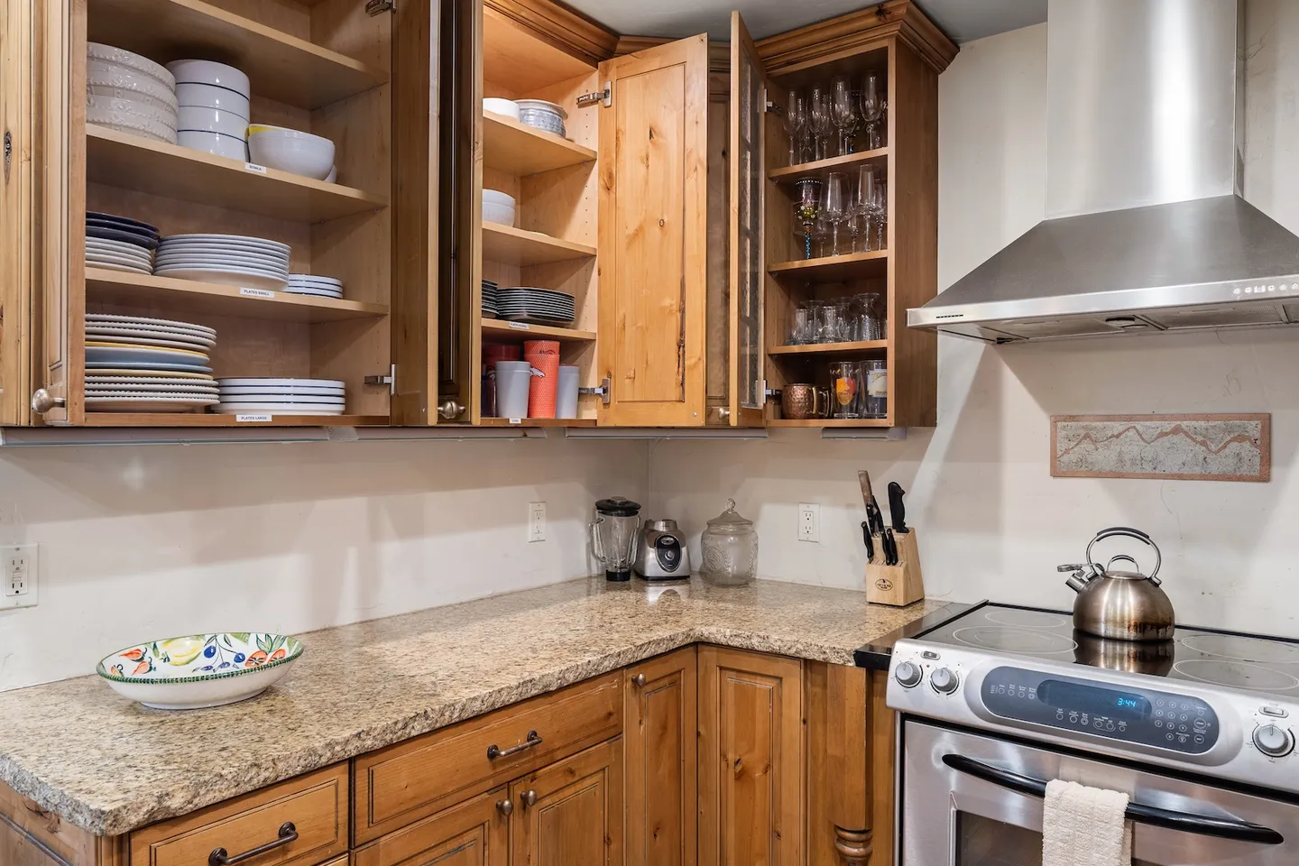 Kitchen counter and open wooden upper cabinets displaying stacked plates, bowls, and glassware alongside a stove and range hood.