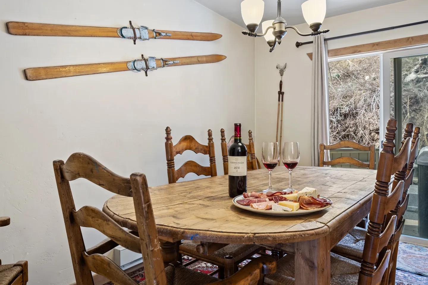 Rustic wooden dining table and chairs set with a bottle of wine, two glasses, and a charcuterie platter beneath vintage skis mounted on the wall.