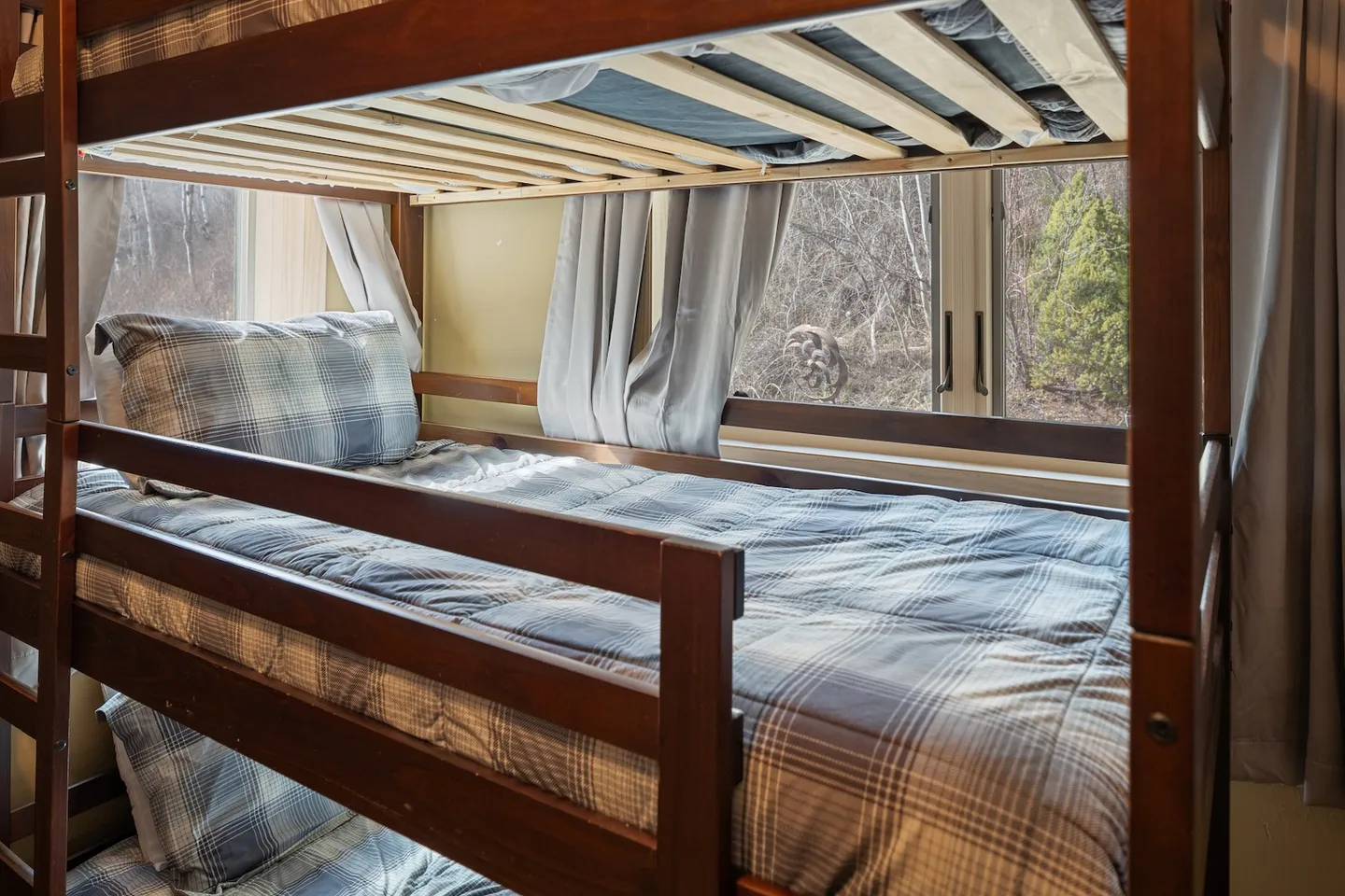 Close-up view of wooden bunk beds fitted with blue and gray plaid bedding next to a window overlooking woods.