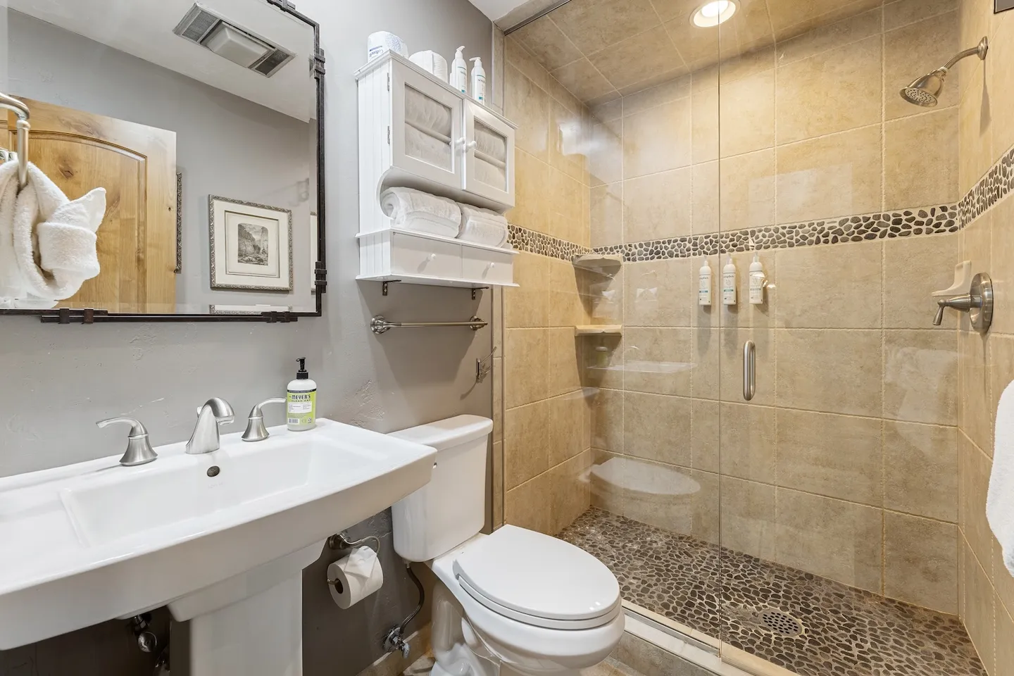 Bathroom featuring a pedestal sink, framed mirror, toilet, and walk-in shower with glass doors and stone tile.