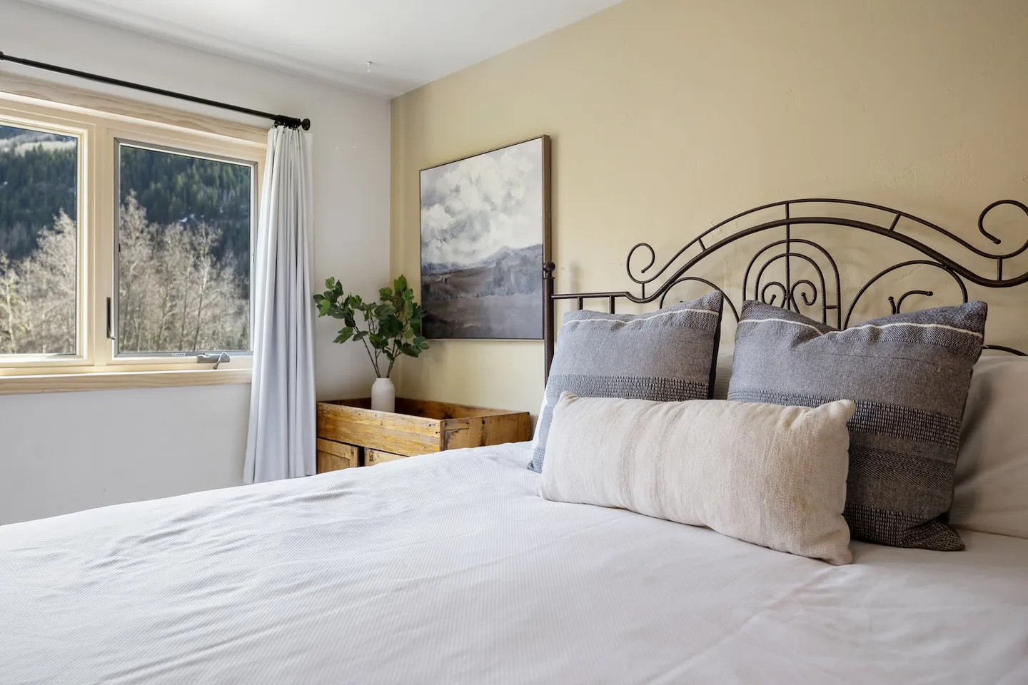 Bedroom with a wrought-iron headboard, made bed, side table, wall art, and a window with views of wooded hills.