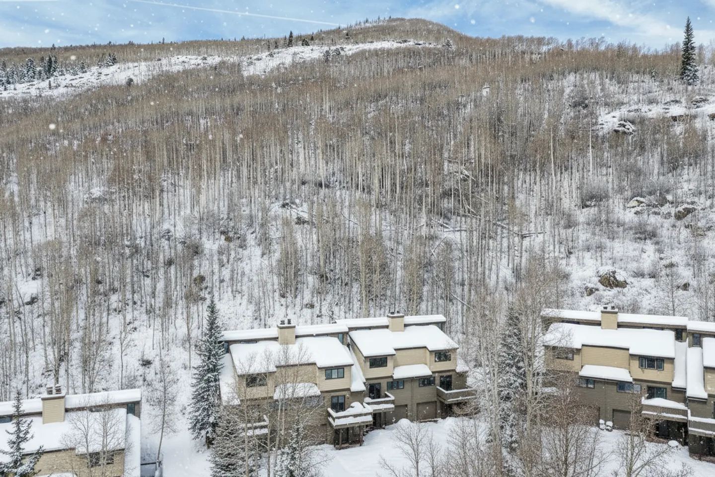Elevated view of multi-story townhomes nestled at the base of a snow-covered hillside with dense bare trees during light snowfall.