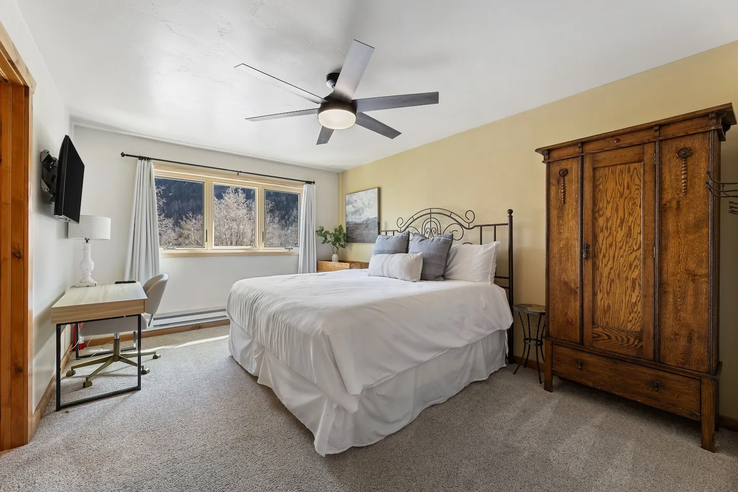 Bedroom with carpeted flooring, a bed with a wrought iron frame, a ceiling fan, a tall wooden armoire, and a small desk with a wall-mounted TV.