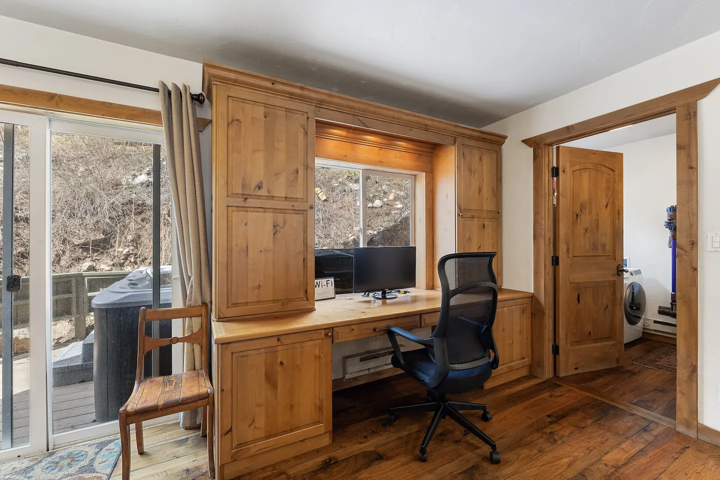 Built-in wooden desk with monitor and mesh office chair next to sliding glass door leading to an outdoor deck.