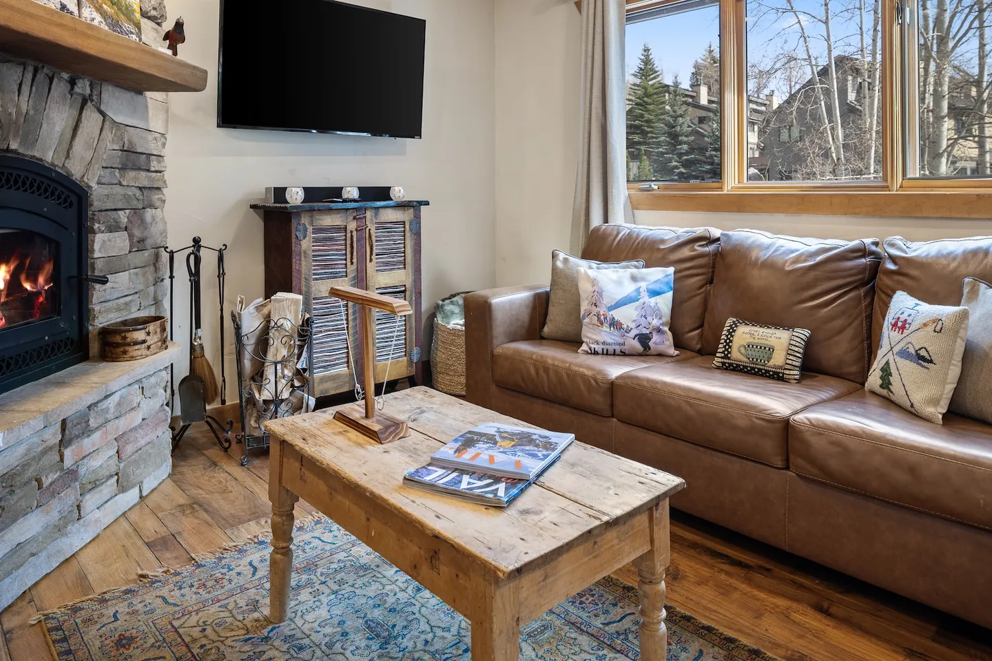 Living room featuring a leather sofa, wooden coffee table, stone fireplace with a fire lit, and a wall-mounted TV.