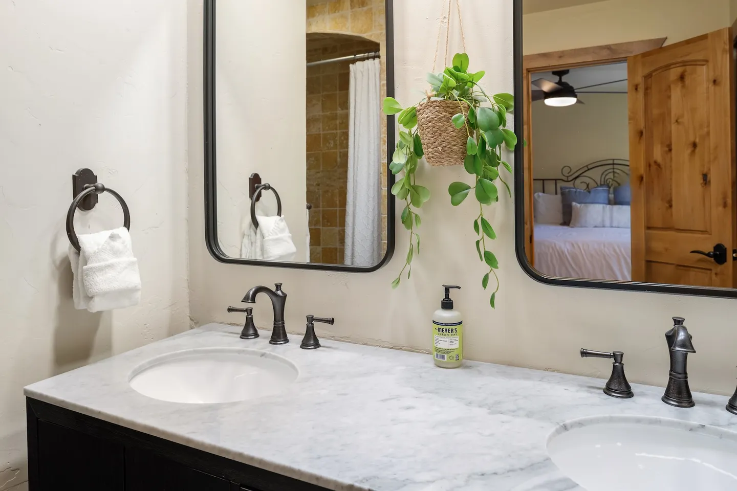 Double sink bathroom vanity with dark fixtures, rounded black-framed mirrors, and a hanging green plant.