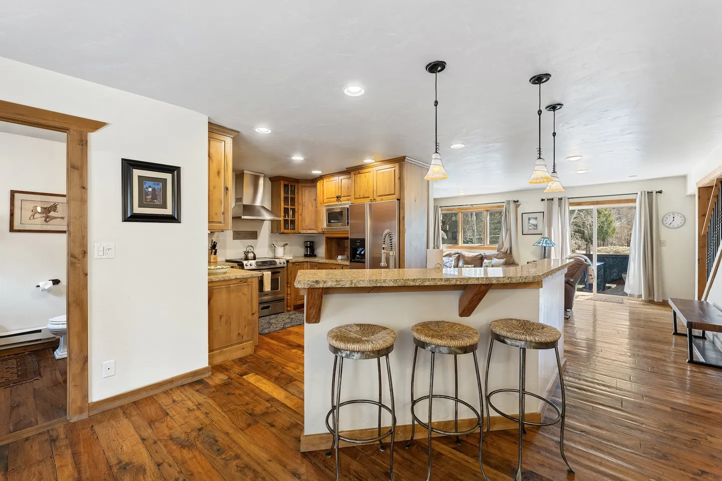 Open-concept kitchen with wood flooring, wood cabinetry, a breakfast bar with three stools, and an adjacent living area.